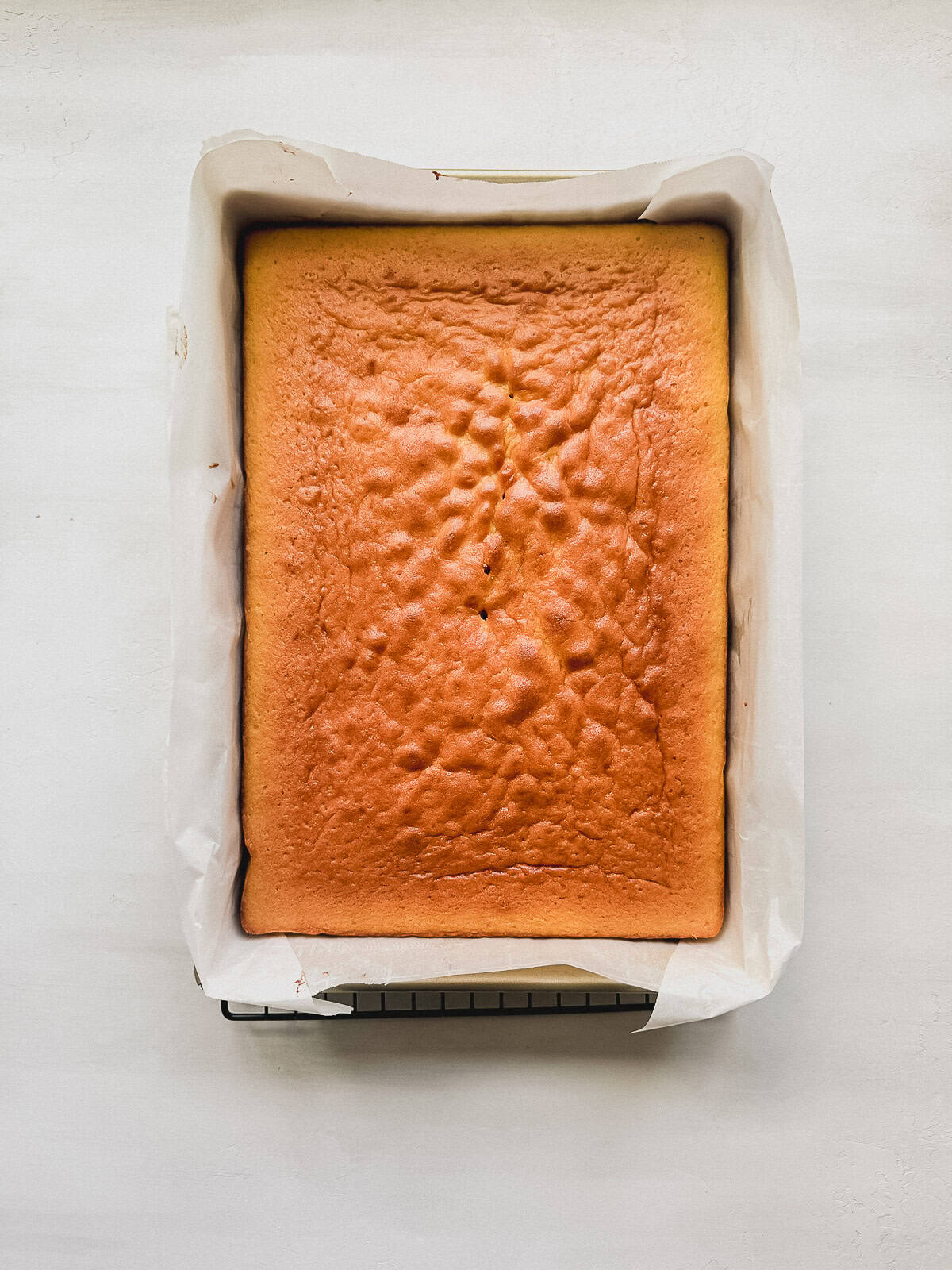 Baked vanilla sheet cake in a 9x13 cake lined with parchment paper on a wire rack.