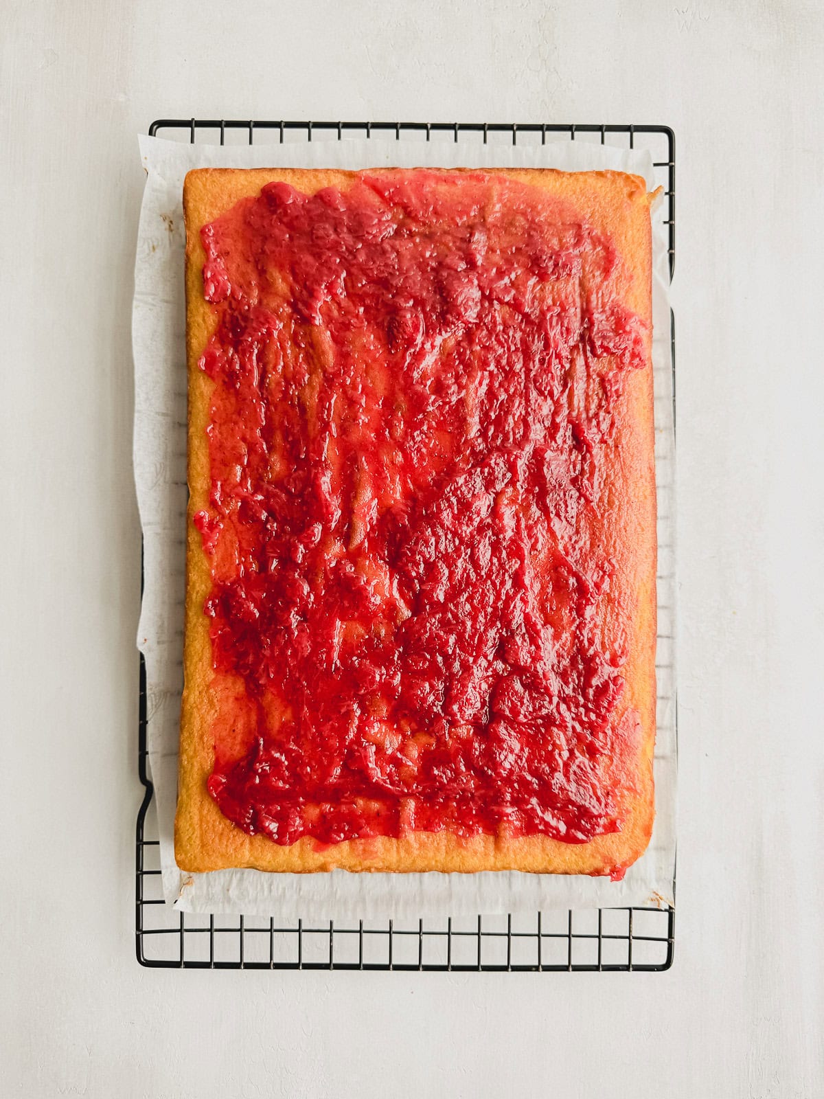 Vanilla cake topped with a layer of strawberry filling on a wire rack lined with parchment paper.